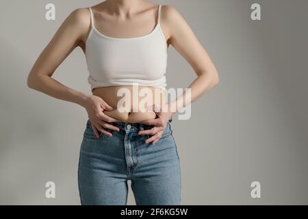 Woman in jeans and white shirt squeezing her belly fat. Woman`s figure closeup raw studio shot in grey background. Dieting and fat loss concept. Stock Photo